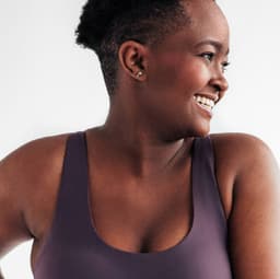In front of a neutral background, woman in sports clothes holding right part of her waistband with both hands, looking and smiling to the left.