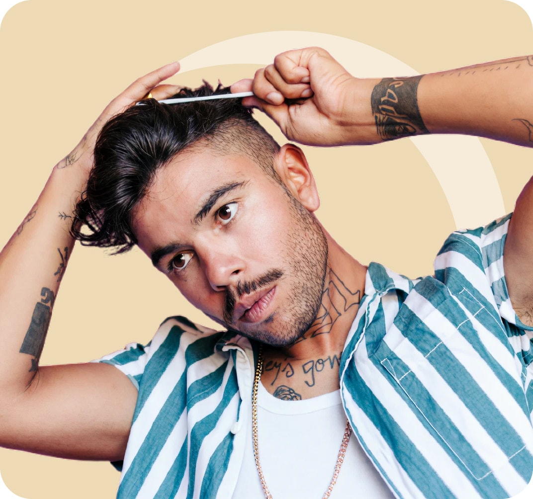 Man with tattoos combing his hair while looking slightly down. He wears a striped shirt over a white tank top and stands in front of a beige background.