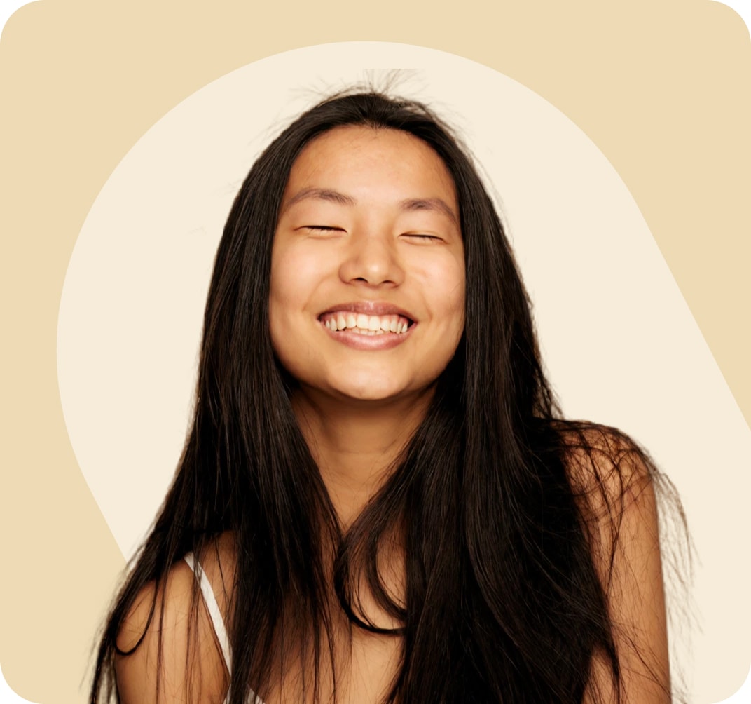Woman with long black hair, smiling with eyes closed in front of a neutral beige background. She is wearing a white tank top with thin straps.