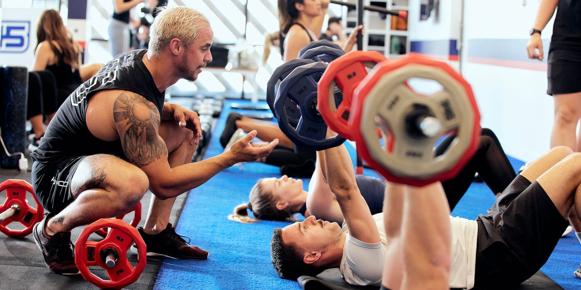 Color photograph of a white male F45 Training personal trainer kneeling down to assist F45 gym members who lay on the floor, raising barbell weights above them. Photo courtesy F45.