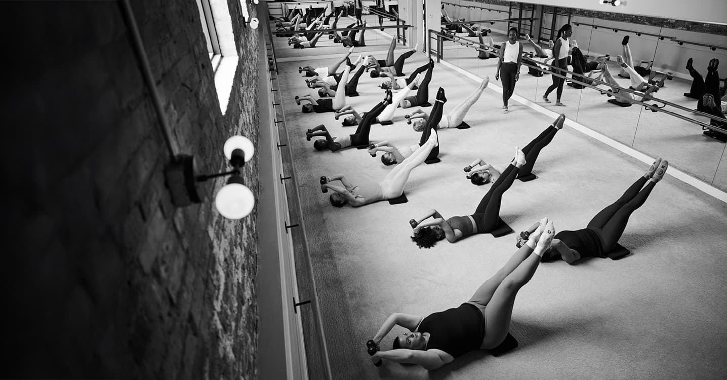 A group fitness class where participants lie on mats lifting their legs and arms with dumbbells, engaging in a core-strengthening exercise.