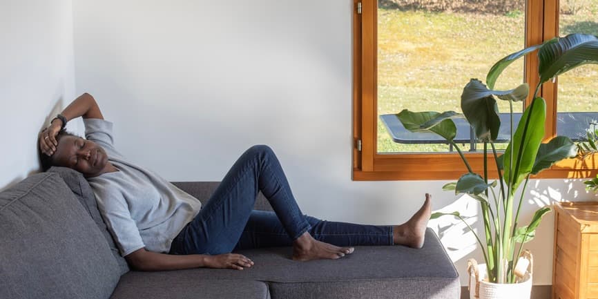 A middle-aged black woman with short hair is resting on a dark grey couch with her eyes closed