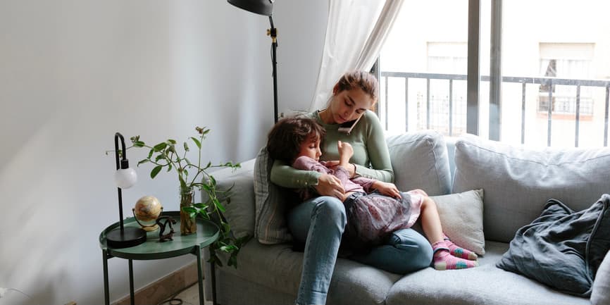 Young brunette woman sitting on the couch in their living room at home, with a 6-year old kid on her lap. She's on the phone doing a consultation with a doctor. The woman is checking the child’s temperature on a thermometer.