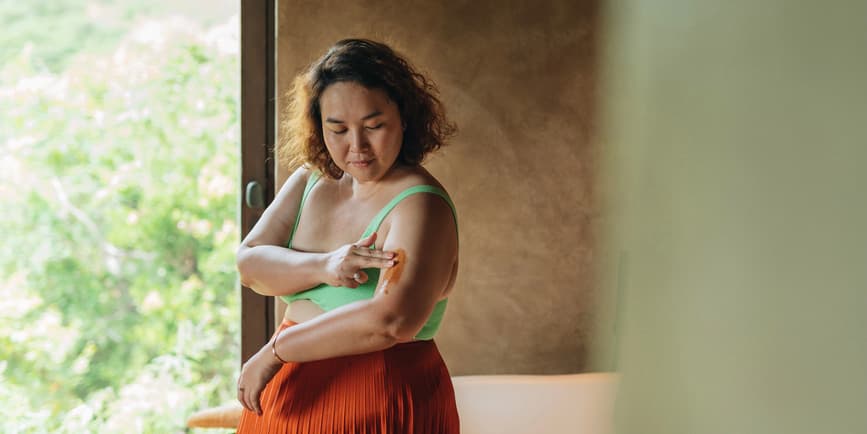 Woman Applying Body Scrub On Hand A curvy woman with brown skin and brown hair wearing a green tank top and orange skirt applies a cream to her upper arm.