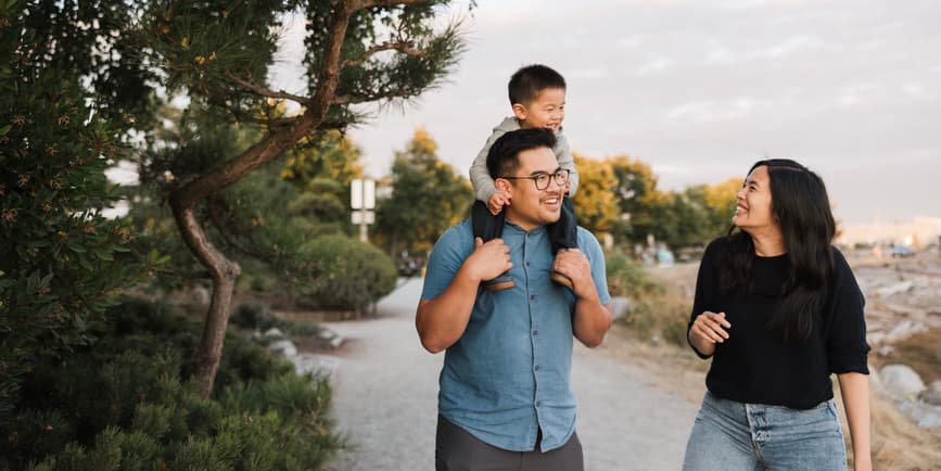 A Latino toddler rides on the shoulders of their father while their mother looks up and laughs. They walk on a broad mountain road, smiling.
