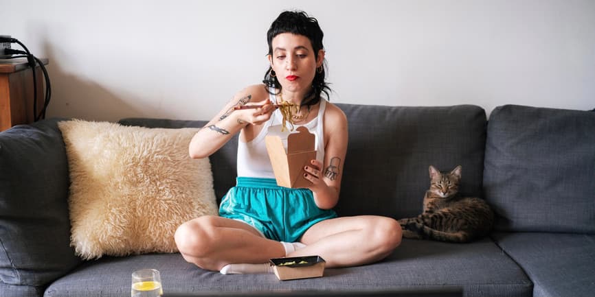 Woman eating on the couch A young white woman with black hair wearing red lipstick eats from a Chinese food container on their couch with their legs crossed, while a brown cat lays nearby.