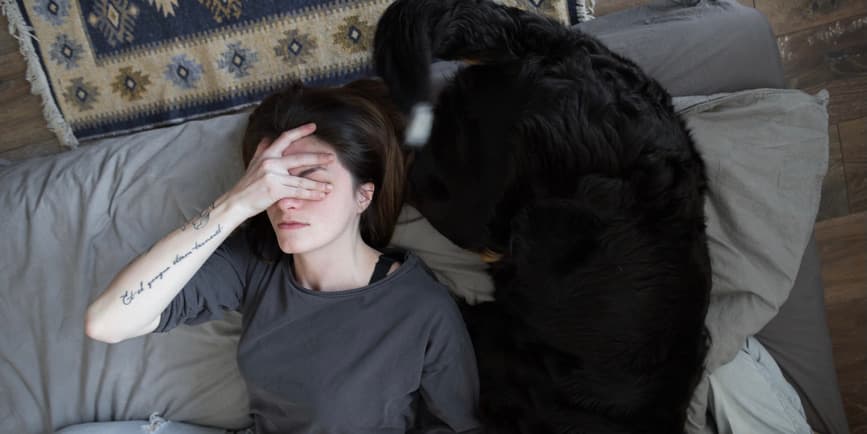 Tired Woman on floor with dog Young white woman with brown hair wearing a gray t-shirt lays on the floor next to a large black dog and covers her eyes with her hand.