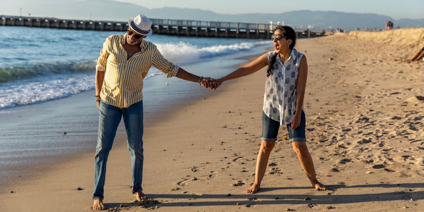 Senior couple on the beach A senior couple with brown/tan skin wearing jeans and summer shirts holds hands while walking on the beach, smiling at each other.