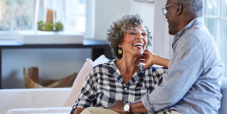 A handsome senior Black couple wearing long shirts sit looking at each other on a couch in a pretty living room.