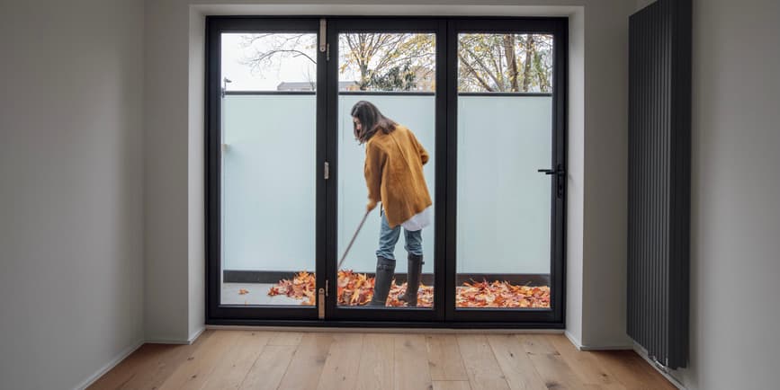 A woman in an orange sweater and jeans rakes leaves on a stone patio, as seen through glass doors from inside.