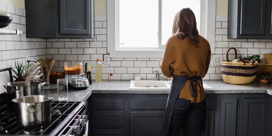 Woman at the sink A white woman with blond hair wearing a brown sweater is viewed from behind washing dishes at a sink in a bright kitchen with black cabinets.