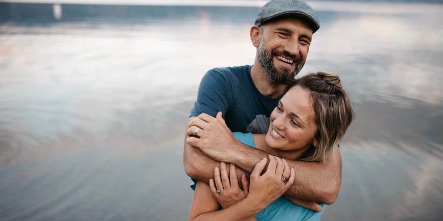 A young white couple wearing t-shirts embrace on a beach with still water behind them. They are both smiling and holding tight.