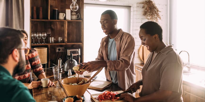 Two young couples smile, talk, and prepare dinner together in a sunlight kitchen full of wood and stone accents.
