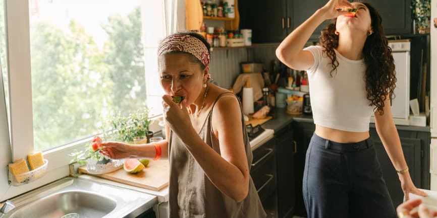Mother and daugther eating fruit in kitchen A Latina mother and adult daughter eating watermelon in their kitchen. The kitchen is full and cluttered, with lots of beautiful colors/