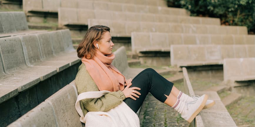 Woman sitting outdoors Woman sitting alone in a relaxed position in outdoor amphitheatre. She is listening to music using earbud headphones and wearing warm clothes.