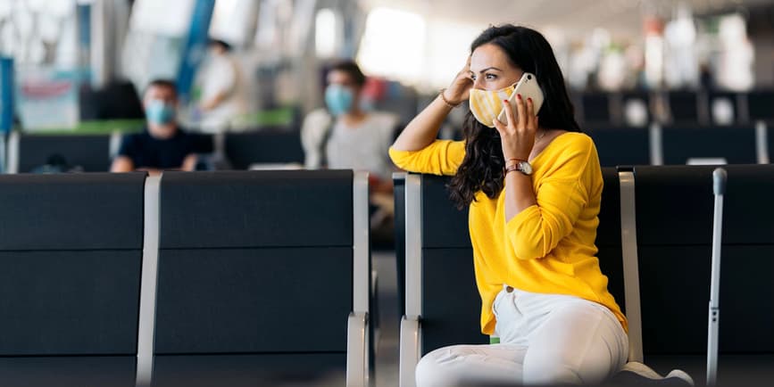 Photo of middle aged white woman wearing a yellow sweater and a face mask, using her phone while she waits for her flight in an airport.
