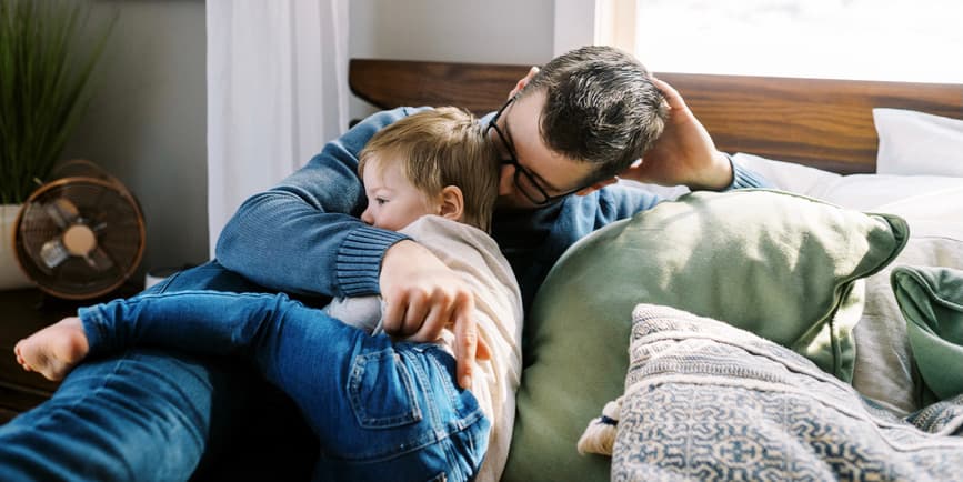 Father and son on bed A young Latino father holds his son while resting on a bed in a bright bedroom.