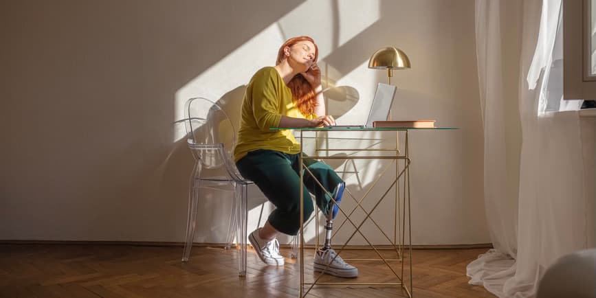 Woman at desk in sun An attractive white disabled woman with red hair and a prosthetic leg enjoys the rays of the sun in her home office.