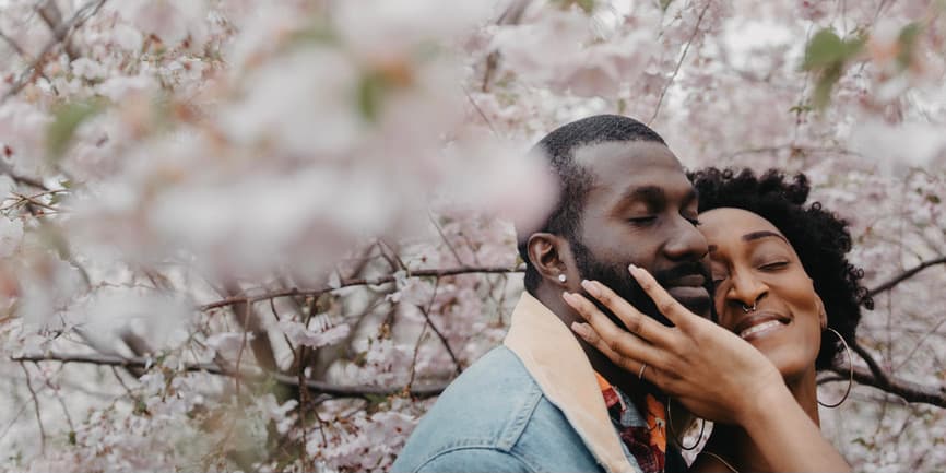 A beautiful young Black couple smile with their cheeks touching as they stand before pink cherry blossom trees.