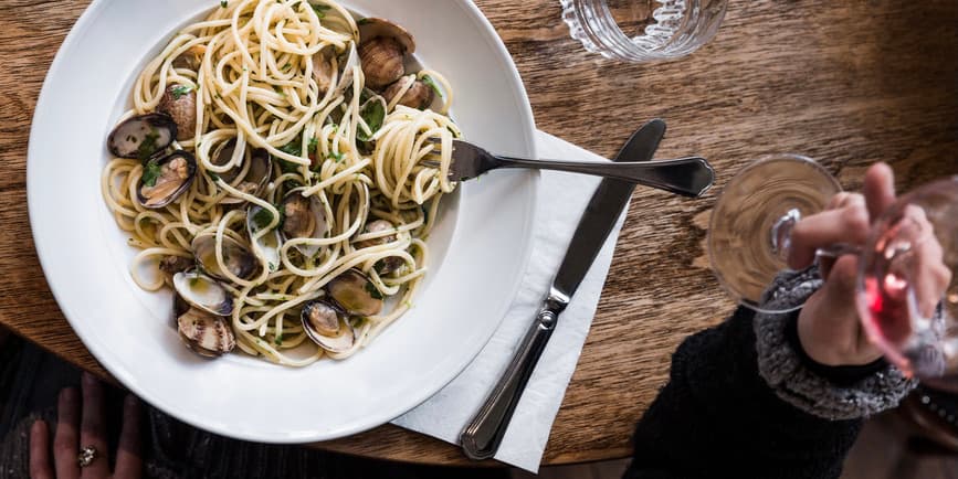 Overhead photo of a woman's hands holding a glass of red wine and a plate of pasta and clams on a wooden surface.