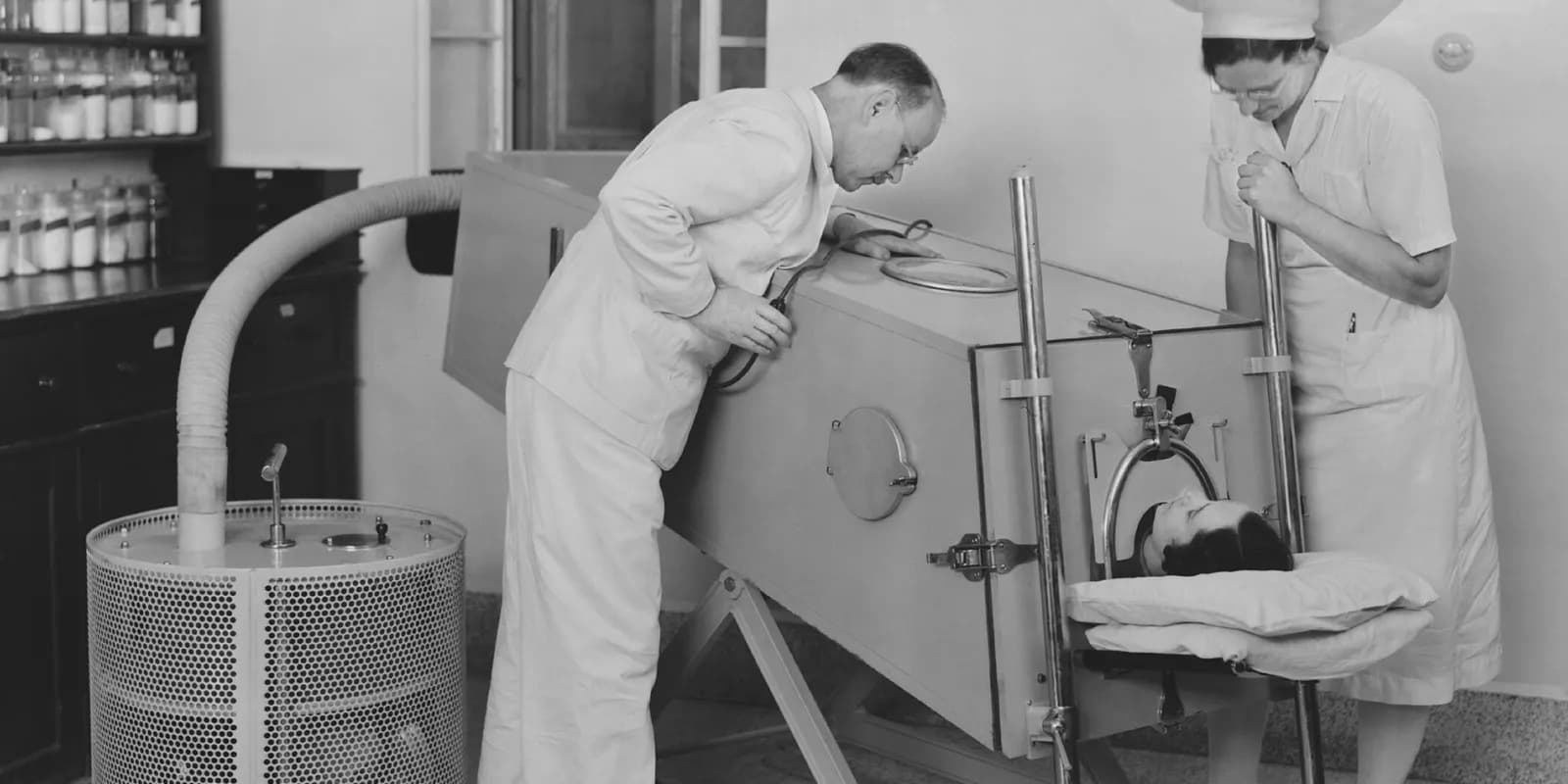 A doctor and nurse treat a polio patient in an iron lung in a black and white photograph.