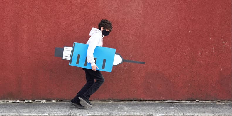 A teenager wearing jeans, a sweatshirt and a medical mask runs against a red building holding a poster cutout of an vaccine needle sign to help people find a vaccine line.