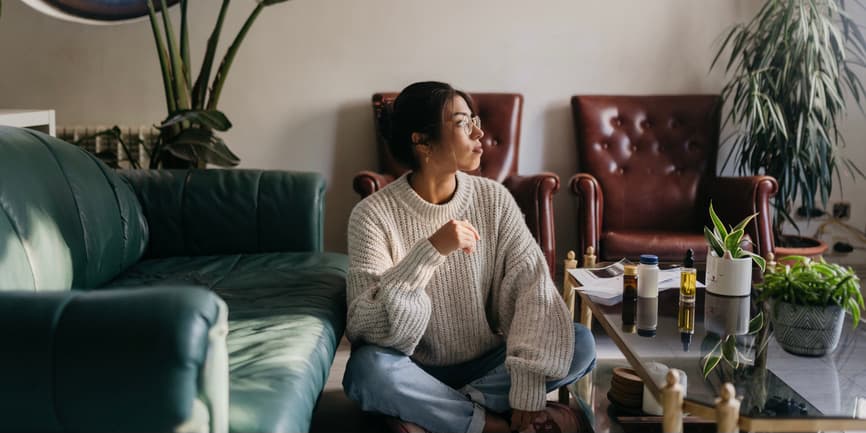 A young Hispanic woman wearing a thick sweater and jeans sits on the floor of her living room, looking out the window, with a bunch of medication bottles on the coffee table next to her.