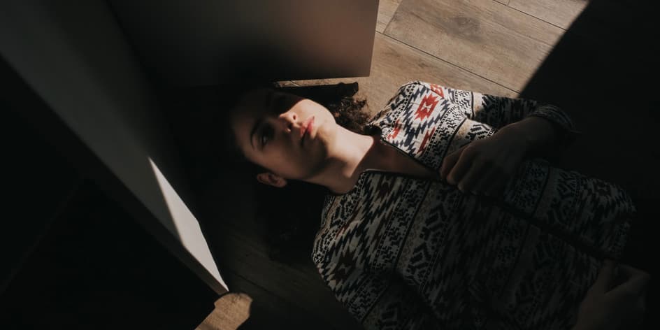 Woman on floor with migraine A. young white woman wearing a colorful cardigan lies on a floor with her eyes closed, half hidden from the sunlight by a half-closed door.