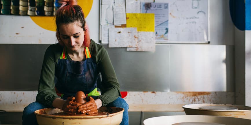 Woman at potter's wheel A young white woman with pink and brown hair tied back wearing a green shirt and blue apron with rainbow ties sits looking down at a potter's wheel as she works clay with her hands.