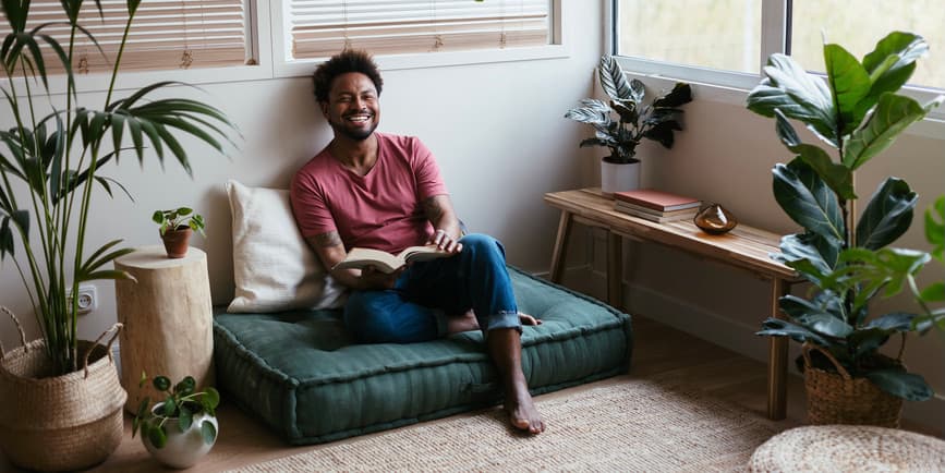 Full body photograph of a young Black man in casual clothes laughing at a joke while sitting on a soft floor pad and reading a book at home in a sunlit room surrounded by plants and wood and rope accents.
