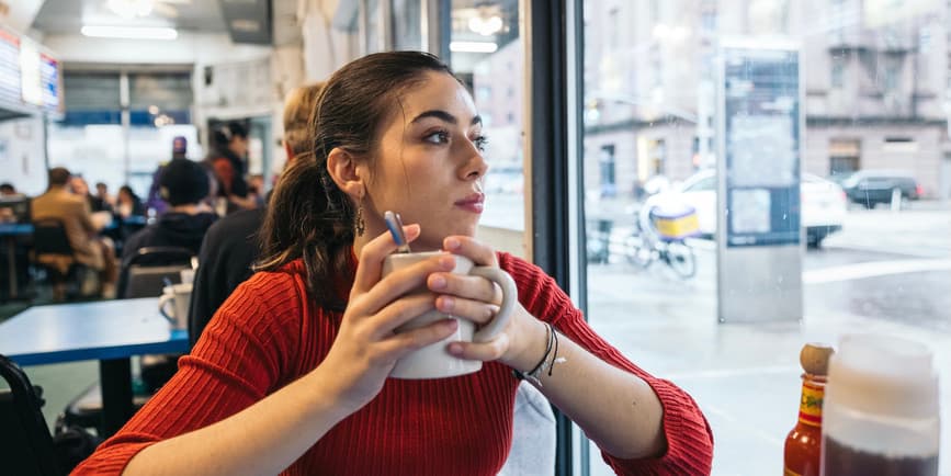 young woman in a diner A young Latina woman wearing a red longsleeved shirt sits in a New York City diner holding a mug of coffee and looking out the window.