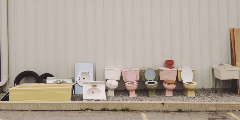 Toilets against a building wall An outdoor photo of a pink exterior wall with discarded toilets in various colors lined up against it.