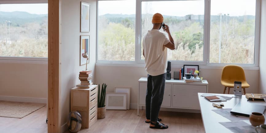 Man at a window A young Black man stands in a modern office with his body looking out a window, away from the camera, talking on a cell phone while gazing out onto mountains of trees.
