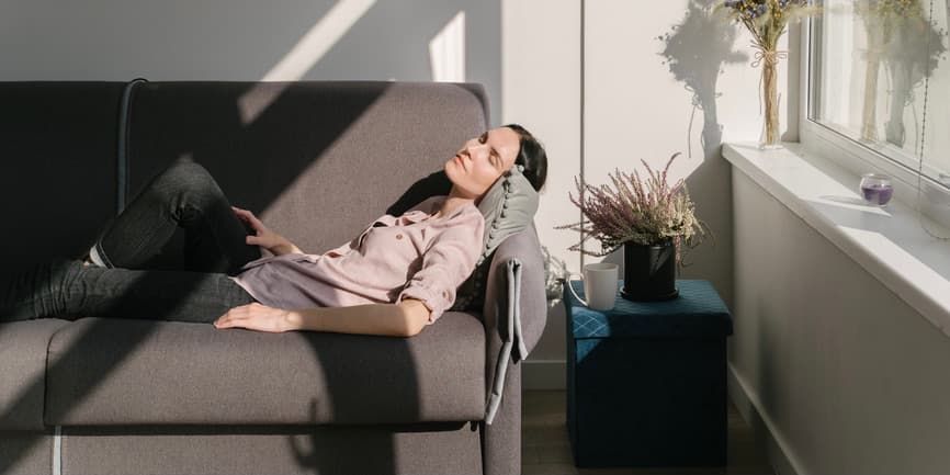 Woman resting on couch A young white woman with long brown hair lays on a gray couch, resting with her eyes closed as sunlight streams onto her from a nearby window.