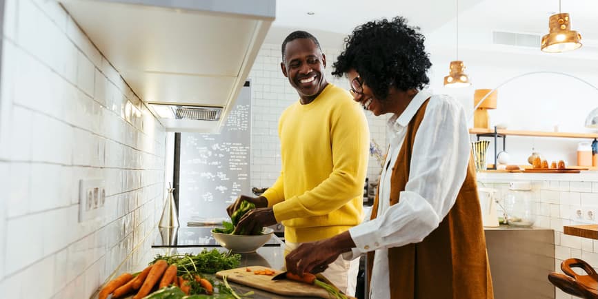 Couple cooking in a kitchen Cheerful Black man and woman in trendy casual clothes smiling and looking at each other while standing near table with fresh vegetables and cutting vegetables for salad in kitchen.