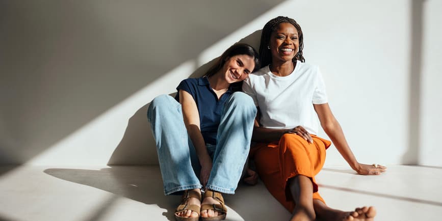 Female friends sitting on the floor Portrait of happy women in trendy clothes spending time together while sitting on floor in bright studio