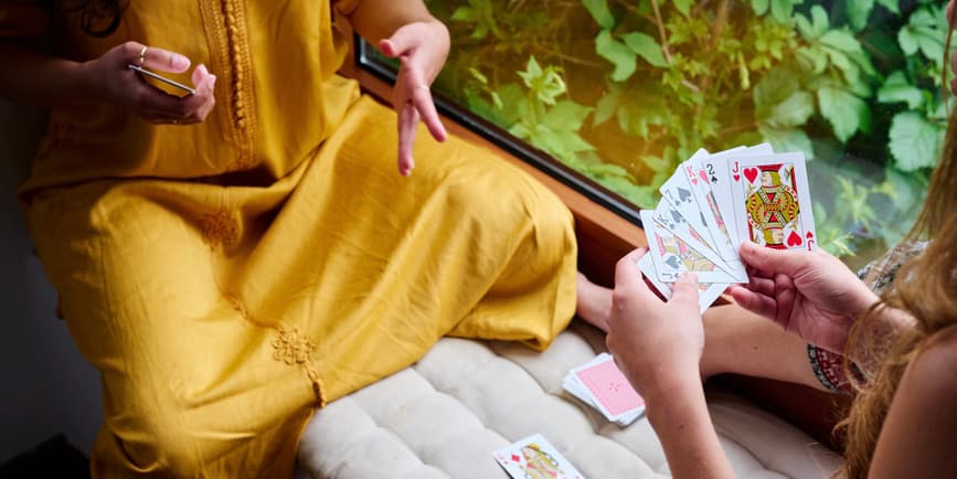 Women playing cards by window Two unrecognizable cropped white girlfriends in spring clothes playing simple card game in a window seat with greenery outside. One explains the rules, the other girl holds a spread of cards in her hand.