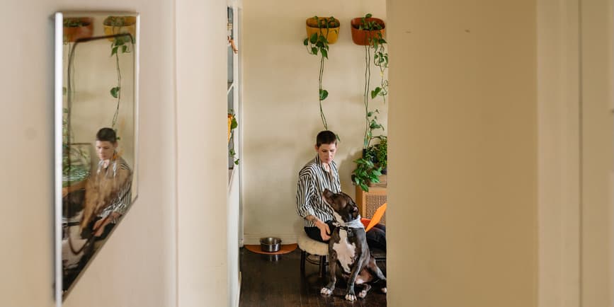 Woman at desk with dog LGBTQ+ woman inside their home working on laptop while petting their large dog who is sitting on the floor next to them. The photo is taken from far away with walls and a doorway framing the shot and part of their reflection shown in a side mirror.