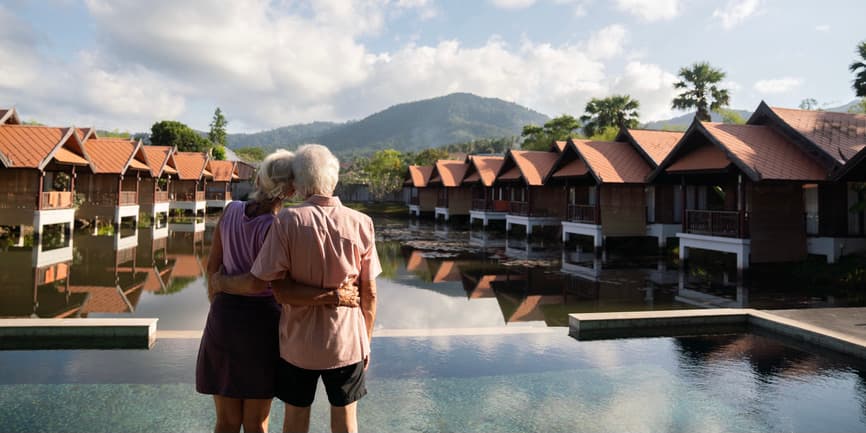 Older adult couple staring at sky View from the back of a senior couple holding onto each other and enjoying a scenic mountain nature view overlooking a hotel pool area.