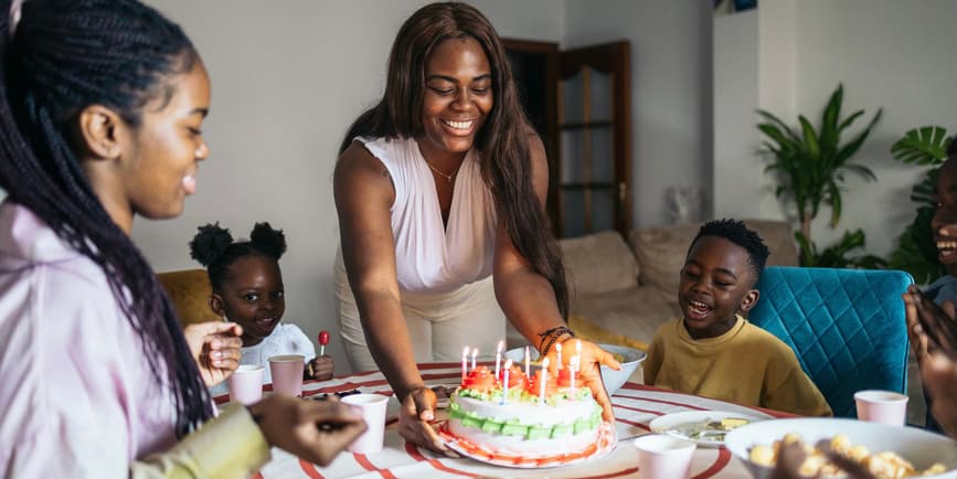 Family birthday party A Black woman in their living room presents a birthday cake with lit candles for her daughter. They are celebrating her birthday at home as a family with her brothers and little cousins. Everyone is happy enjoying the moment together