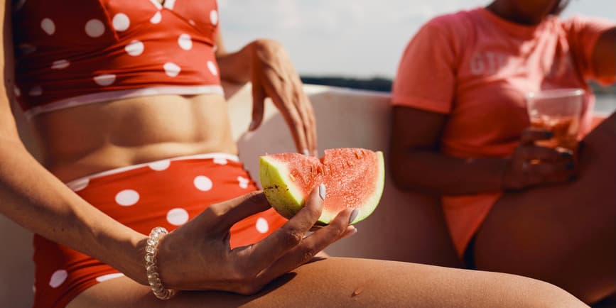 Women eating watermelon in the sun A closeup shot of two friends sitting in the sun, one wearing a polka dot tankini bathing suit, holding a slice of watermelon. We cannot see their faces.