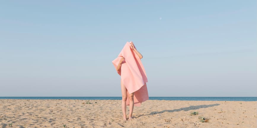Woman covered in towel alone on beach A sandy beach with a pale blue sky behind. At center, a white person with their face and body obscured by a pink beach towel covering their head and most of their body.