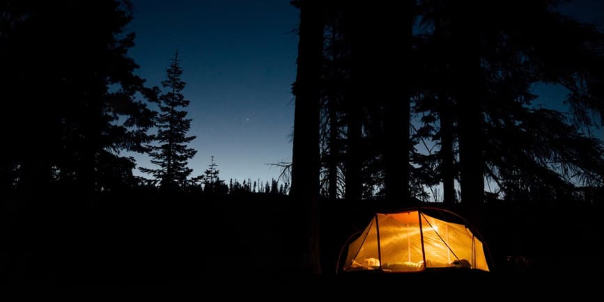 Forest at night with tent A glowing orange tent in the forest at night.