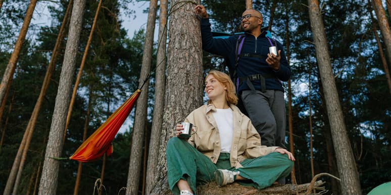 Couple camping A young Black man with a bald head wearing and a young white woman with red hair wear camping clothes and drink coffee from metal mugs at their woodland campsite, smiling as they look at something off camera.