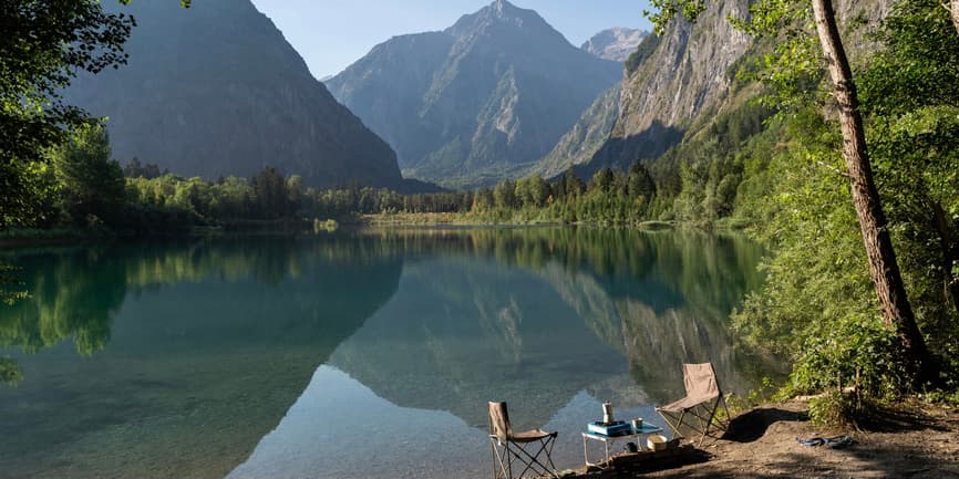 An alpine lake and mountains A broad view of an alpine lake in the mountains, with camping chairs, table, gas stove and coffee maker sitting at the edge and no people in sight.