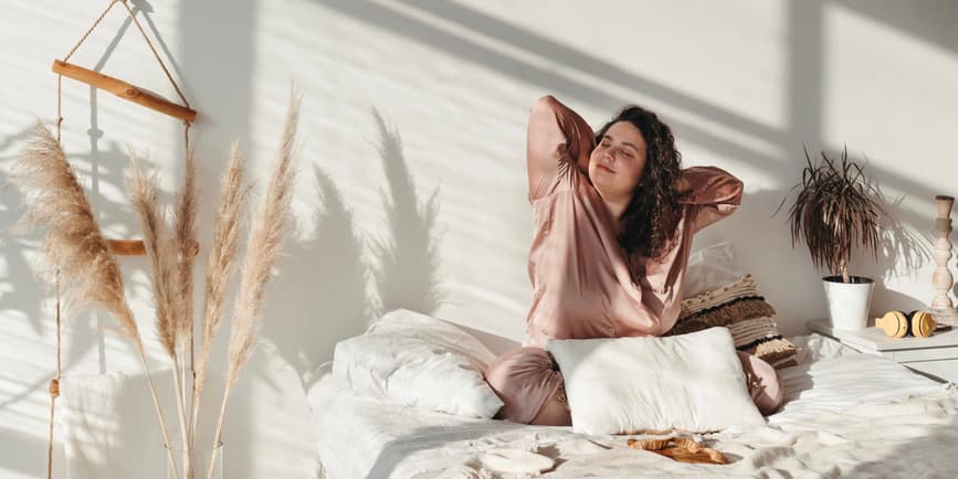 Awakening young adult woman in pastel silk pajamas stretches after sleep and enjoys sunny morning in cozy home environment.