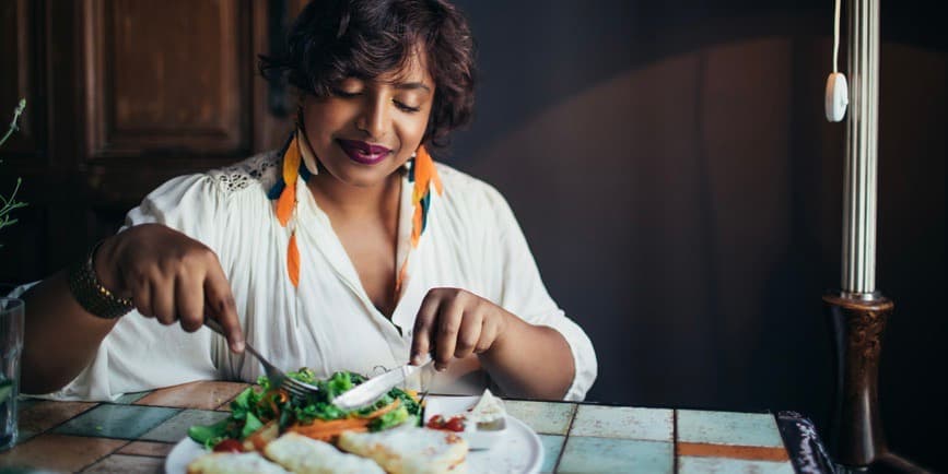 A beautiful Indian woman wearing a white shirt and long feathered earrings sits at a restaurant table and smiles as she cuts into a colorful dish.