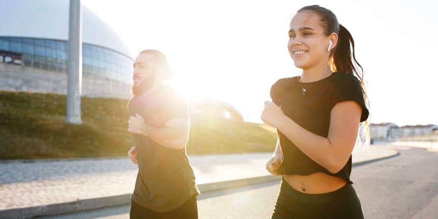 Couple jogging in sunset Cheerful young woman in sportswear smiling and listening to music while running with boyfriend on city street during fitness workout at sundown