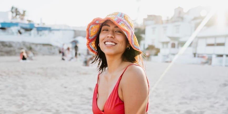 Woman at the beach A young Spanish woman wearing a colorful hat and red bikini smiles joyfully at a busy beach during a sunny day, capturing the essence of summer fun.