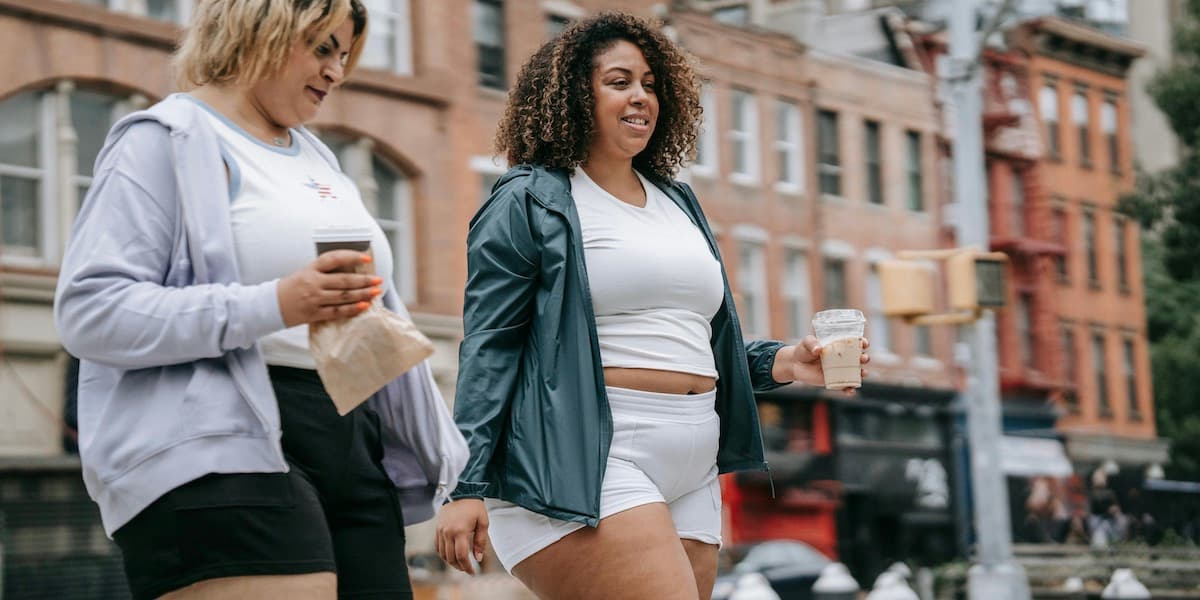 Two female friends with larger bodies walk in New York City in workout clothes after jogging, carrying coffees while smiling and talking.